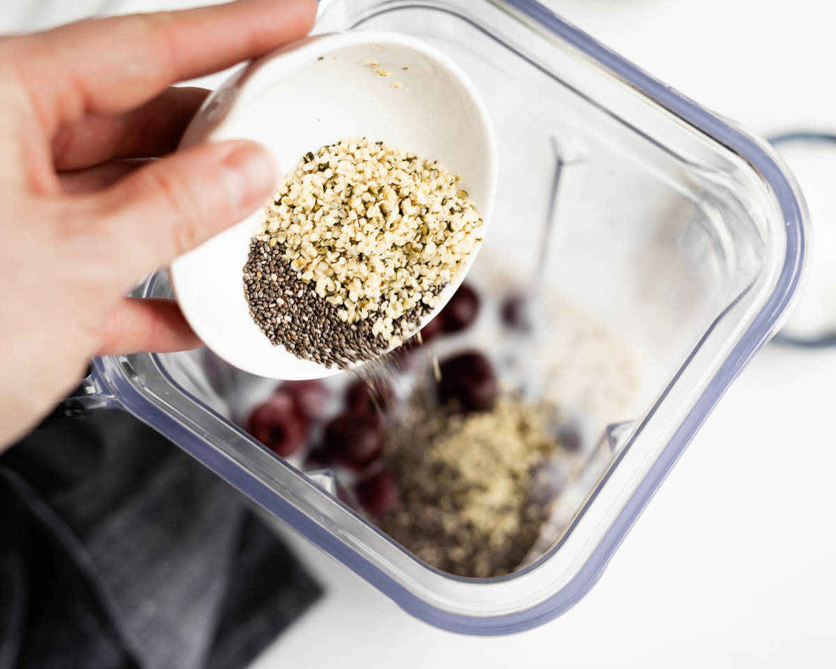 pouring a white bowl of chia seeds and hemp hearts into a blender container.