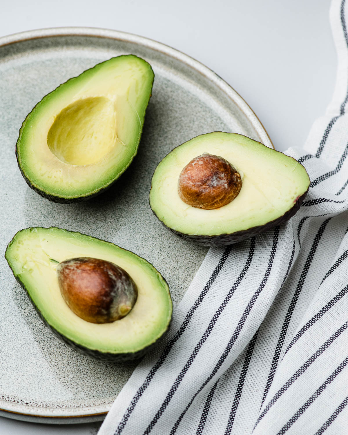 3 halved avocados on a stone plate next to a striped tea towel.