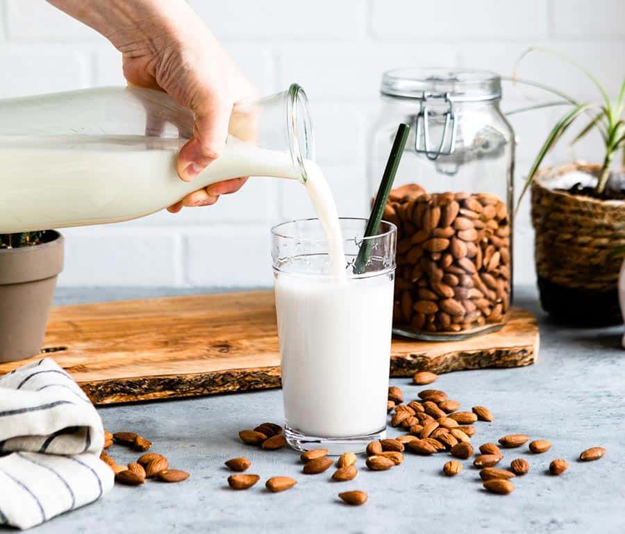 pouring a plant-based milk into a glass with a green straw on a counter surrounded by raw almonds.