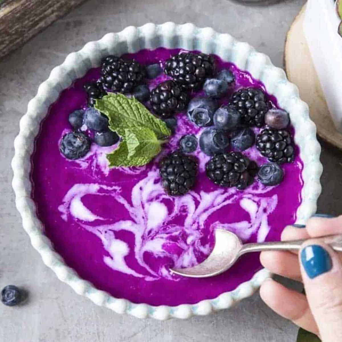 white scalloped bowl with purple dragon fruit smoothie in it topped with blueberries, blackberries, coconut whipped cream and a spoon.