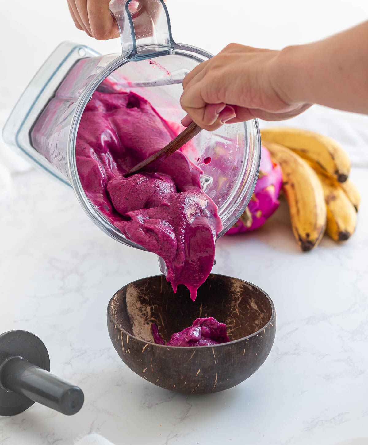 scooping purple smoothie out of a blender container and into a coconut bowl with a wooden spoon.