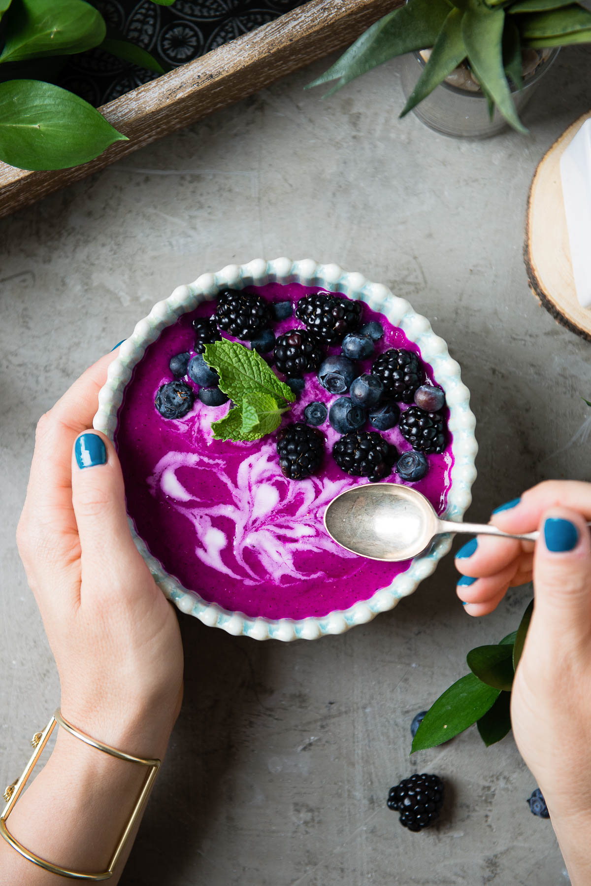 2 hands with blue nail polish dipping a spoon into a dragon fruit smoothie bowl topped with berries and fresh mint.