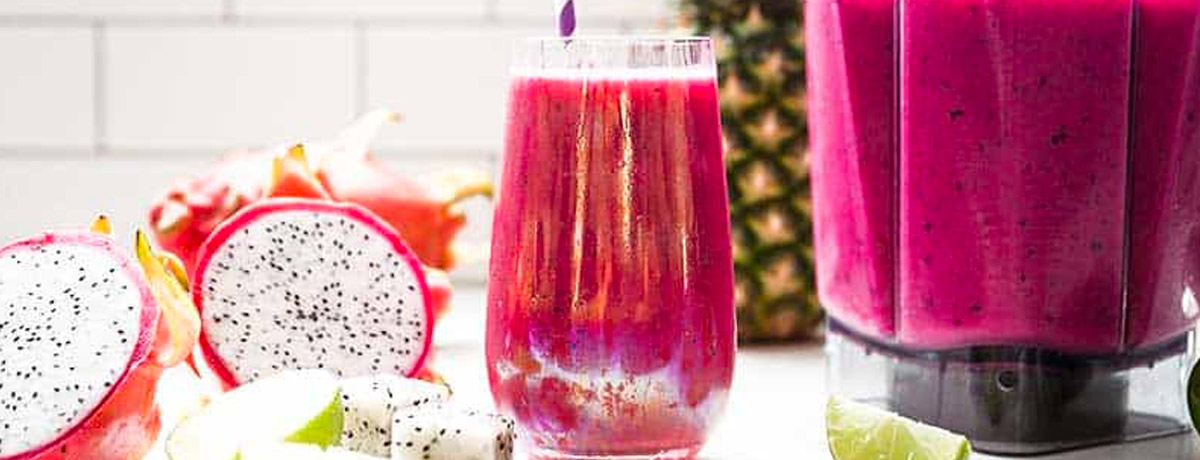 bright pink smoothie in glass with a straw and a blender pitcher filled with the same smoothie and a cross section of a dragon fruit