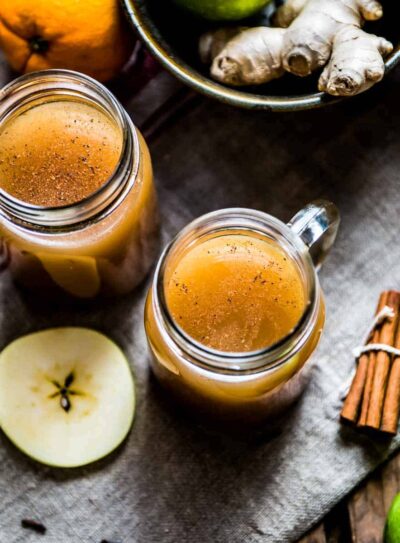 Two glass mugs of crockpot apple cider surrounded by fresh ingredients.