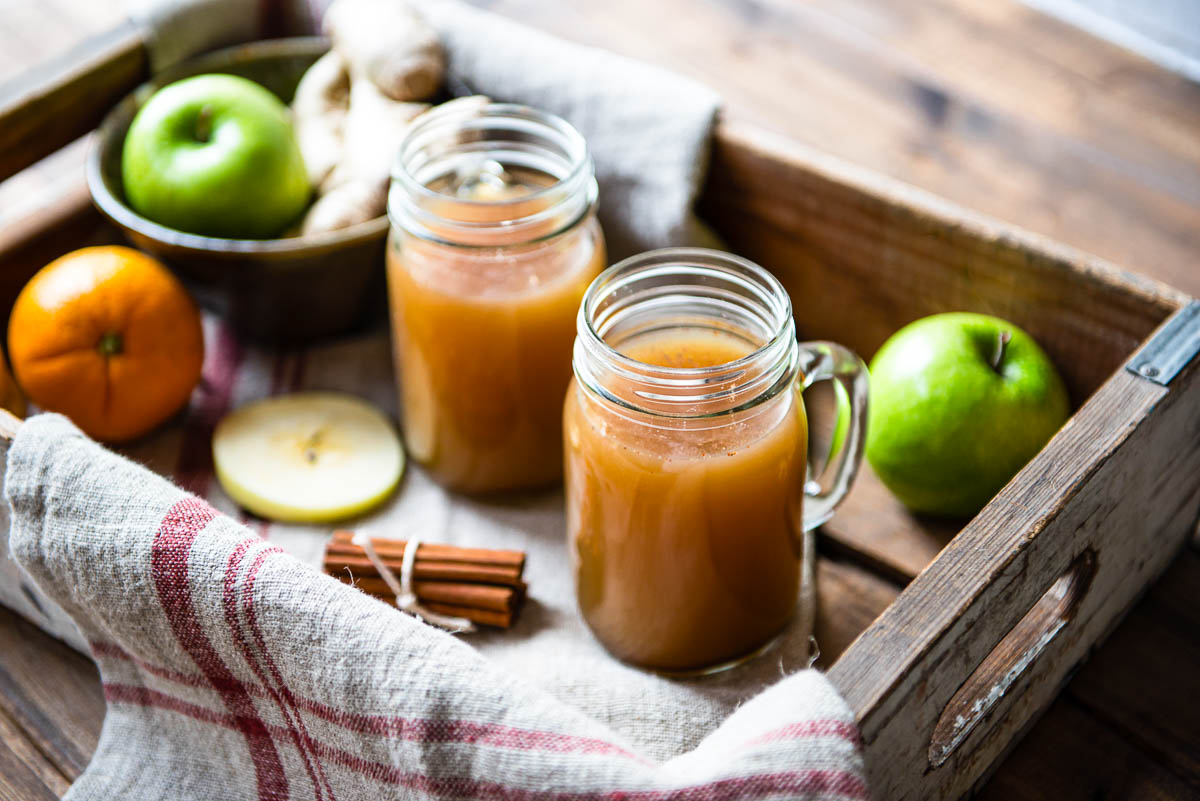 2 glass mugs of hot apple cider in a wooden crate with fresh apples, an orange and a bundle of cinnamon sticks.