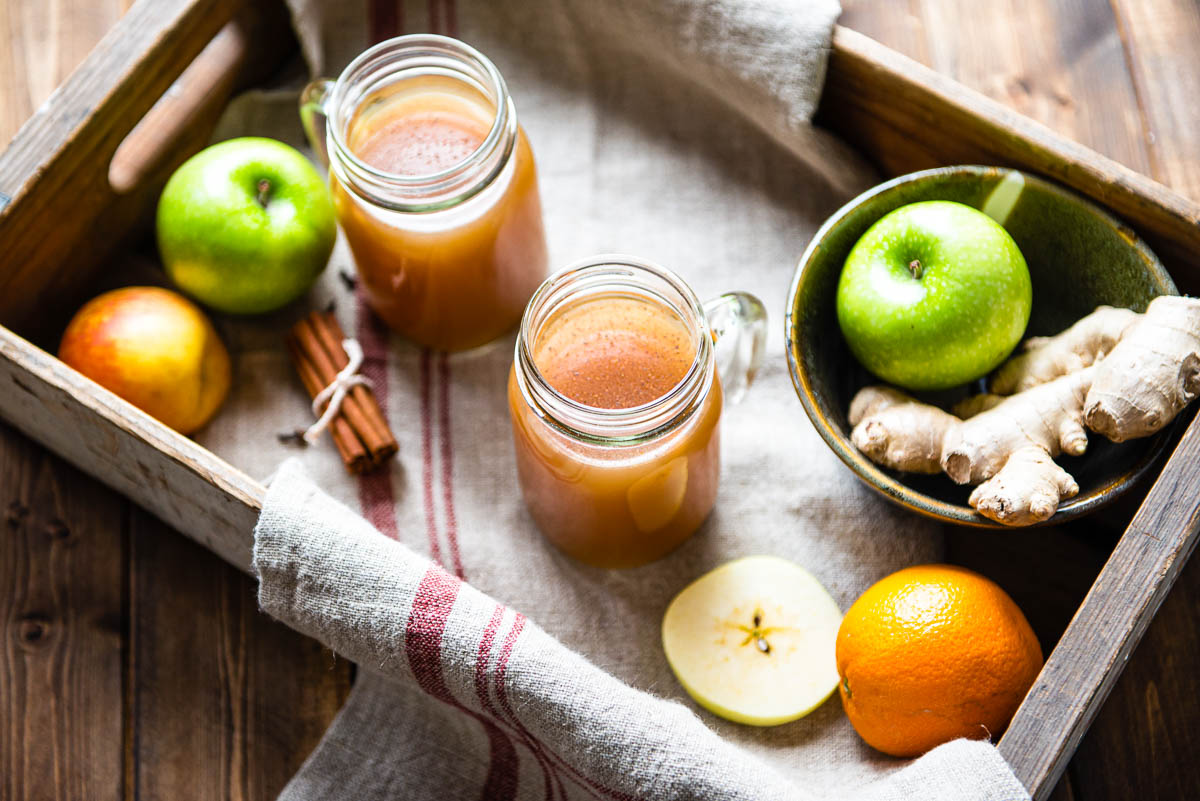 2 glass jars of hot apple cider in a wooden crate next to whole apples, an orange, ginger root and a bundle of cinnamon sticks. 
