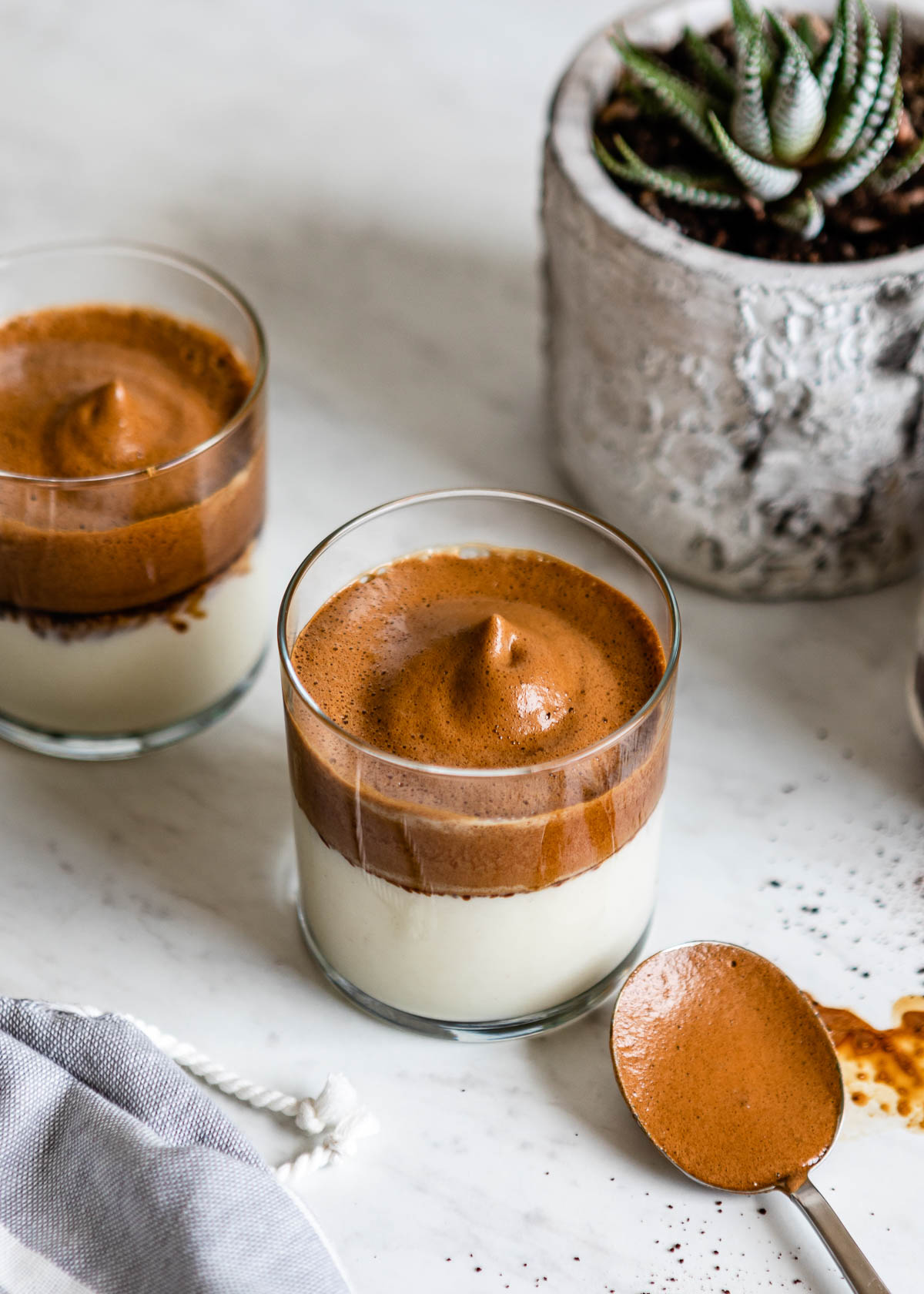 2 cups of whipped coffee next to a spoon and a fake plant.