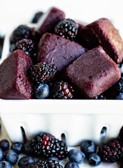 Close-up of bright, purple elderberry smoothie cubes in a white container, surrounded by fresh blueberries and elderberries.