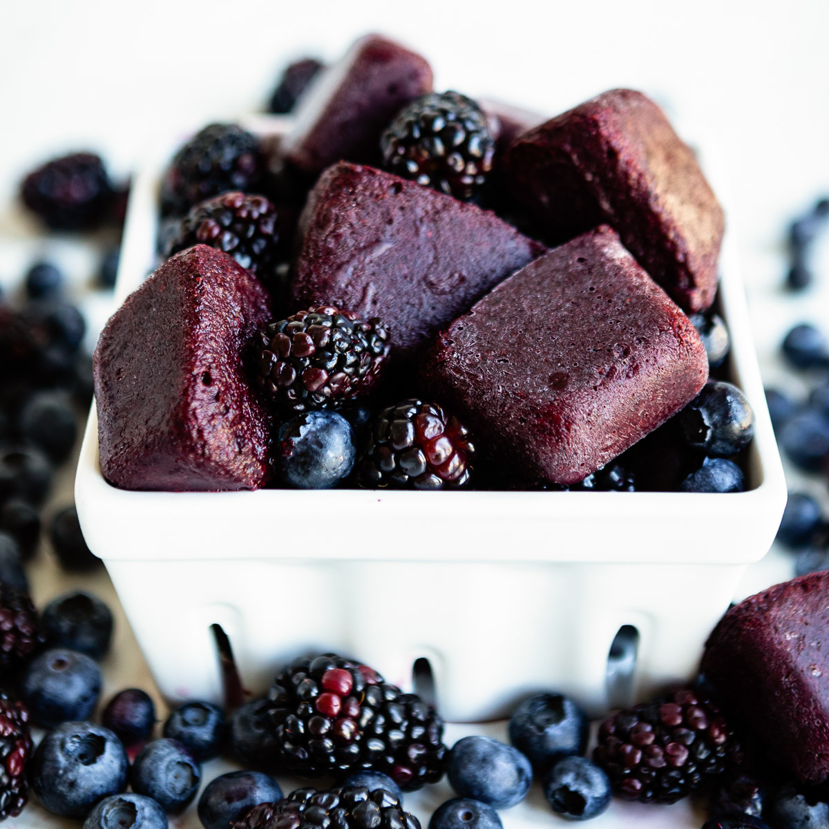 Close-up of bright, purple elderberry smoothie cubes in a white container, surrounded by fresh blueberries and elderberries.