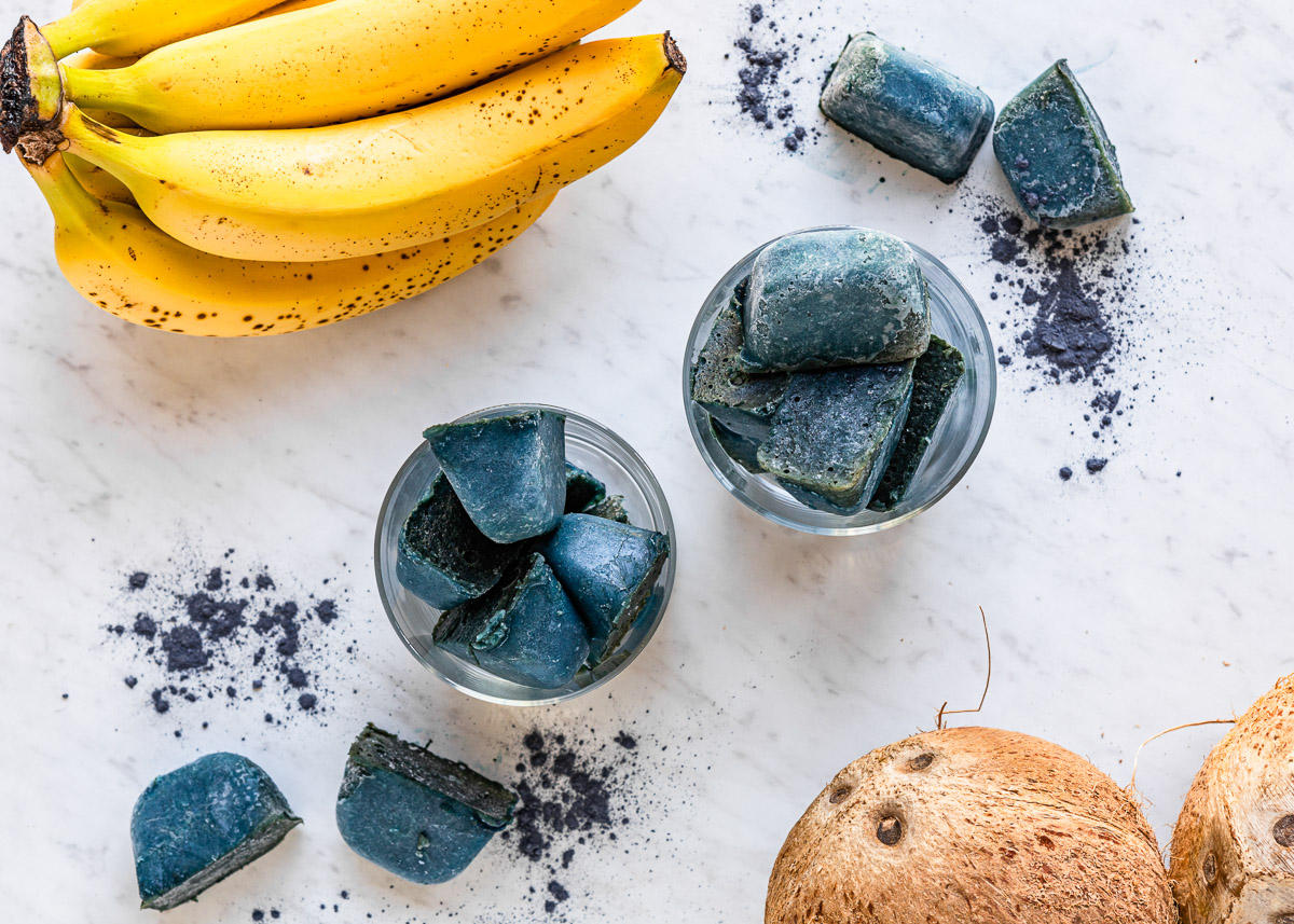 Two glass jars filled with natural electrolyte smoothie cubes, next to fresh bananas and a whole coconut.