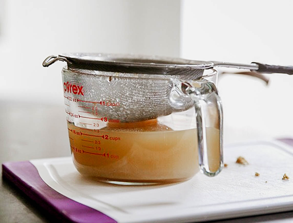 glass measuring cup with brownish liquid in it and a strainer sitting on top.