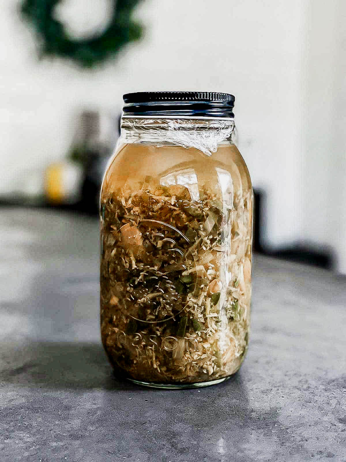 a large mason jar full of ingredients and liquid, wrapped in plastic wrap and a tight fitting black lid