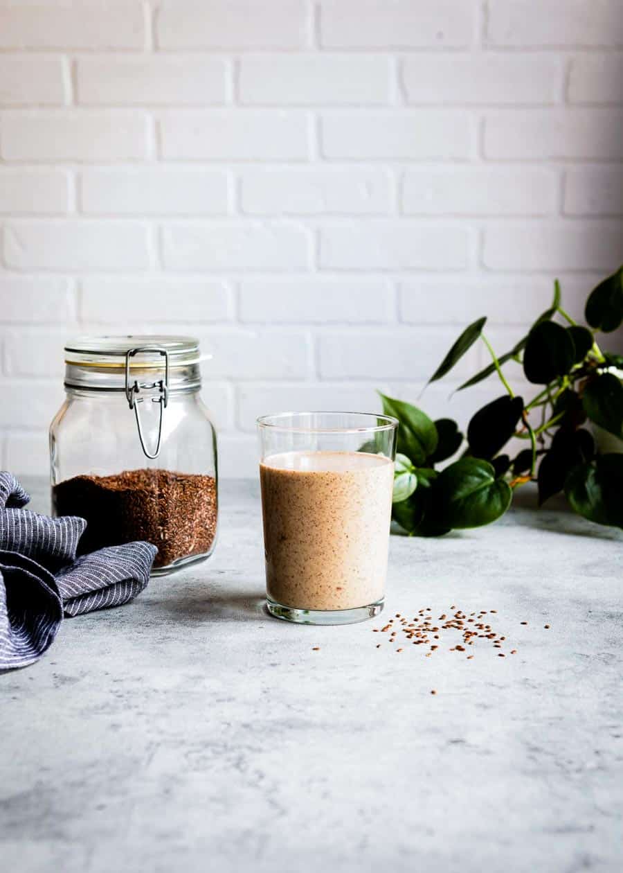 light brown smoothie in a glass cup next to a jar of flaxseed.