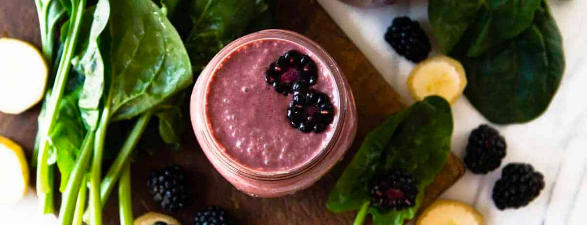 purple smoothie in a jar with fresh blackberries on top and leafy greens in the background