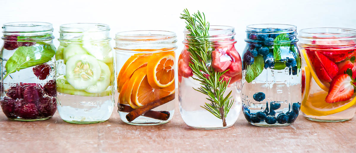 6 Mason jars filled with detox water in a row with berries, oranges, rosemary and grapefruit.