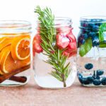 3 glass jars of fruit-infused water with ingredients like oranges, cinnamon sticks, rosemary, strawberries, blueberries and basil.