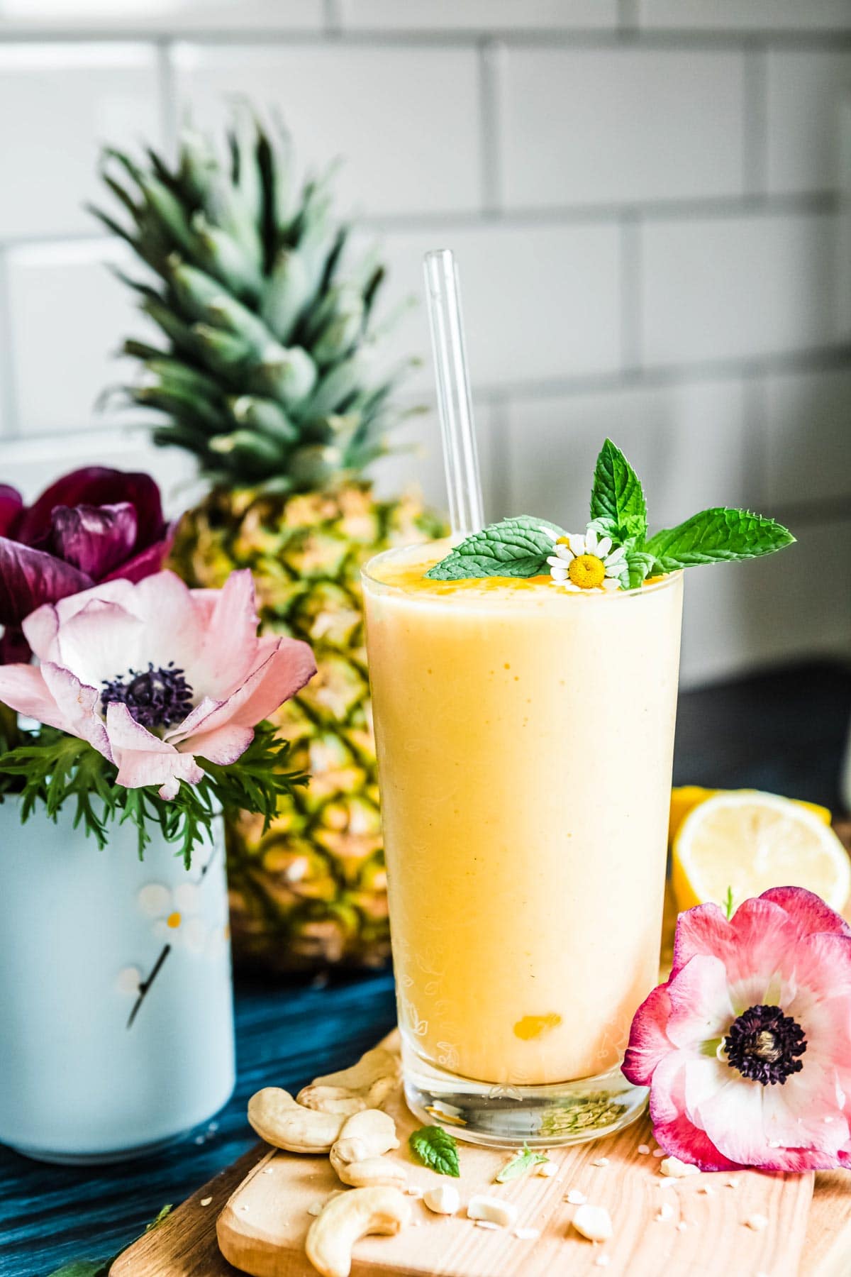 yellow fruit smoothie recipe in a glass with a glass straw and topped with mint, net to fresh flowers and fruit.