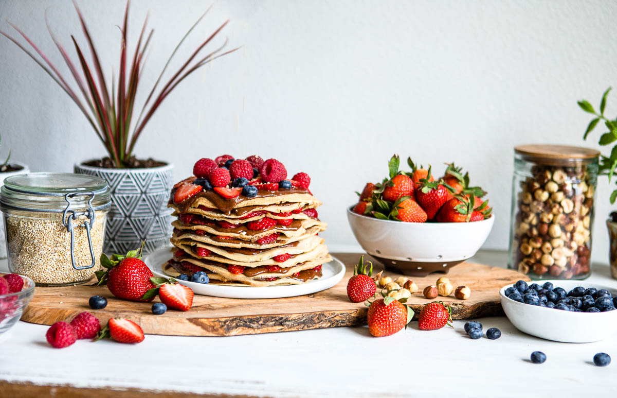 vegan crepes stacked on a white plate surrounded by fresh fruit