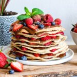 A white plate of layered gluten-free crepes filled with hazelnut chocolate spread and fresh berries.