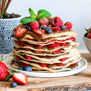 A white plate of layered gluten-free crepes filled with hazelnut chocolate spread and fresh berries.