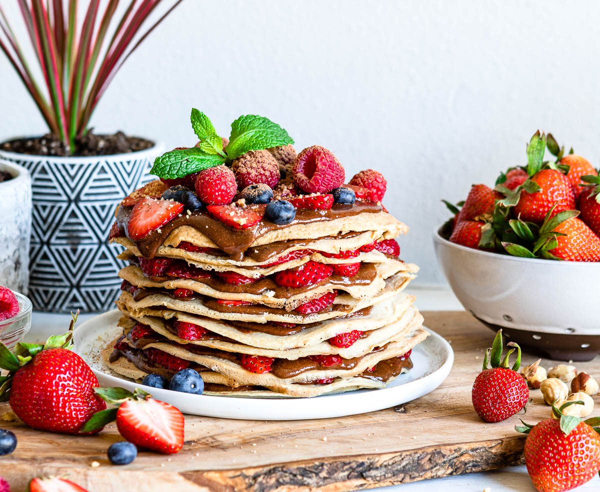 stack of vegan crepes with chocolate hazelnut spread in-between the layers. Fresh raspberries and strawberries top and surround the plate.