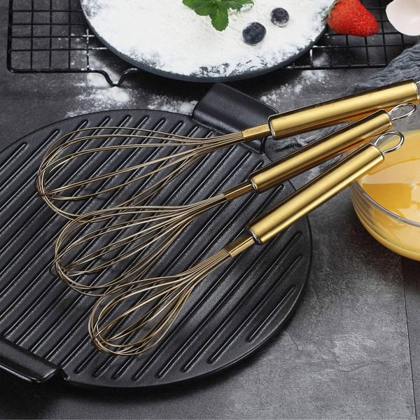 Set of four gold-handled whisks on a black grill pan for cooking.