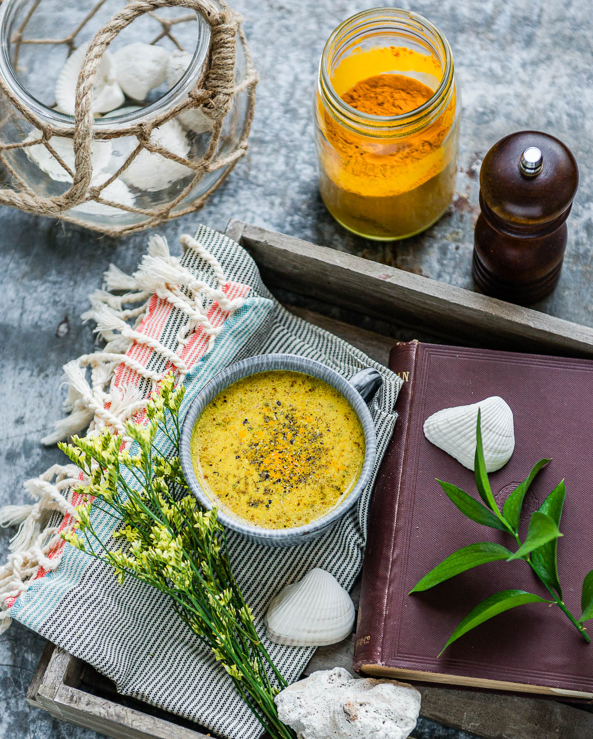 mug of turmeric milk sitting on a tea towel in a wooden tray next to an old book, with cracked black pepper and a jar of ground turmeric sitting above it.