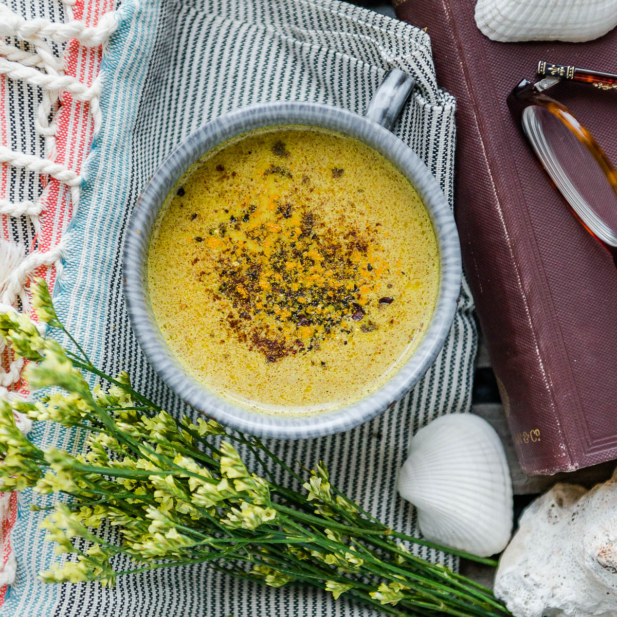 mug of golden milk topped with ground turmeric and cracked black pepper, sitting on a striped tea towel next to some herbs, shells and an old book on a wooden tray. 