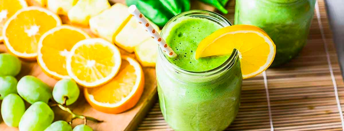green smoothie in a jar with an orange wedge on it and fresh grapes, oranges, and bananas in the background