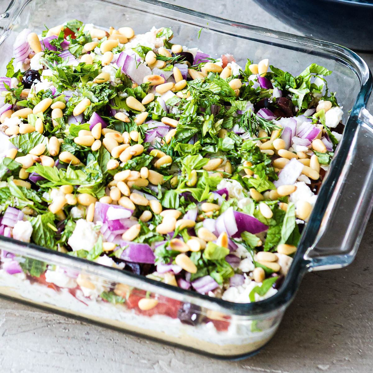 hummus, tomatoes, olives, feta, red onion, parlsey and pinenuts layered in a glass baking dish.