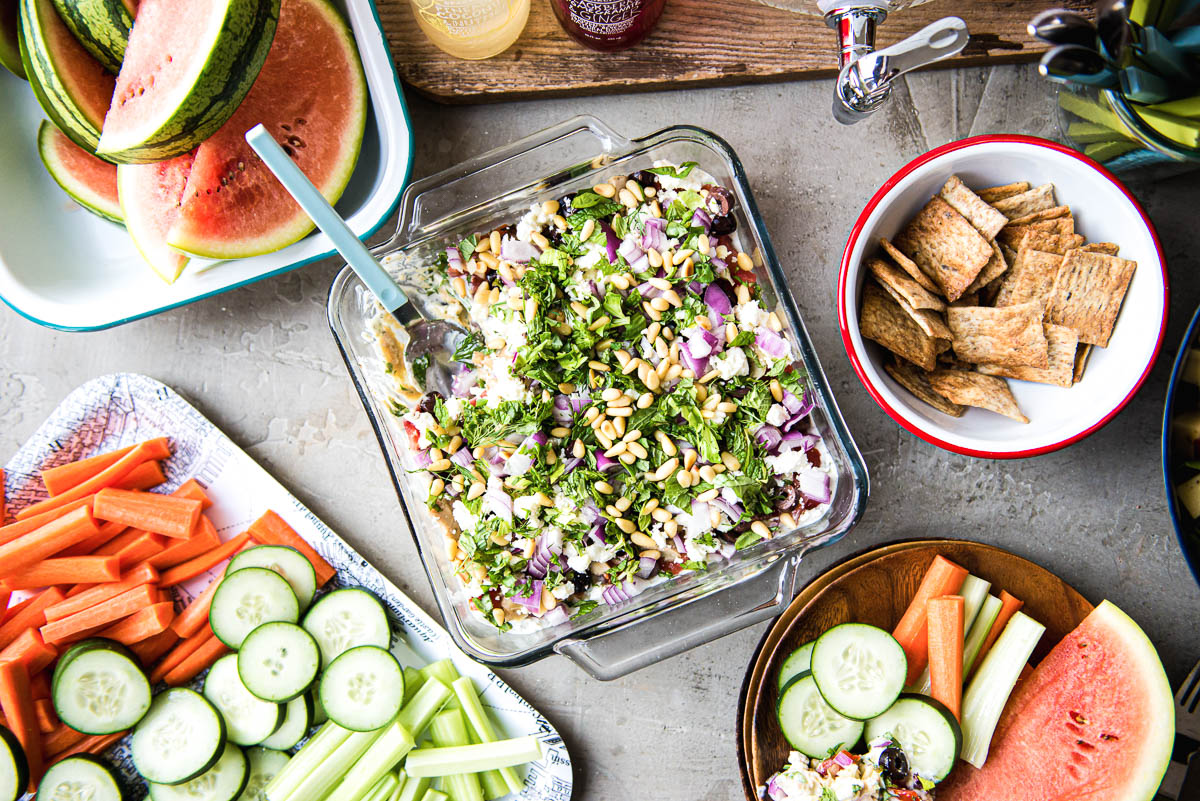 overhead photo of several appetizers including a Greek layered dip, crackers, veggie tray and watermelon slices.