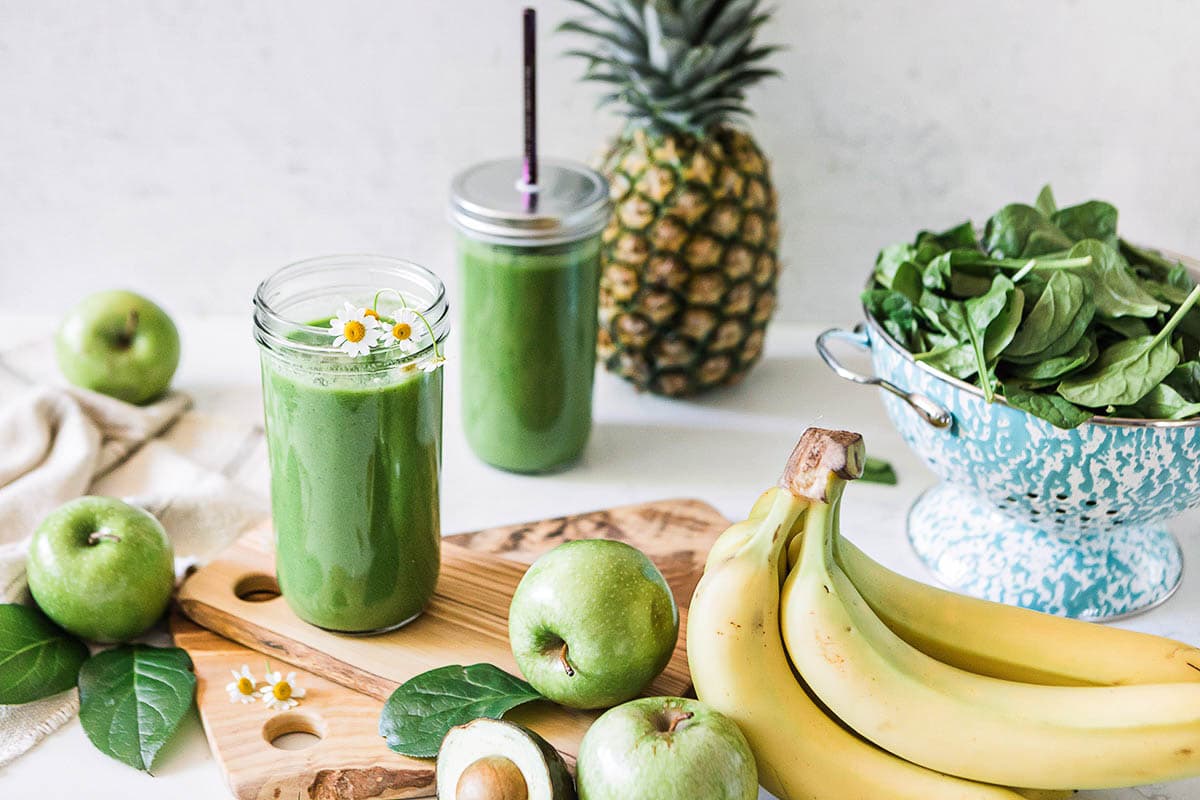 Two jars of green apple smoothie surrounded by fresh spinach, bananas, pineapple, avocado, and green apples and displayed with woodgrain cutting board.