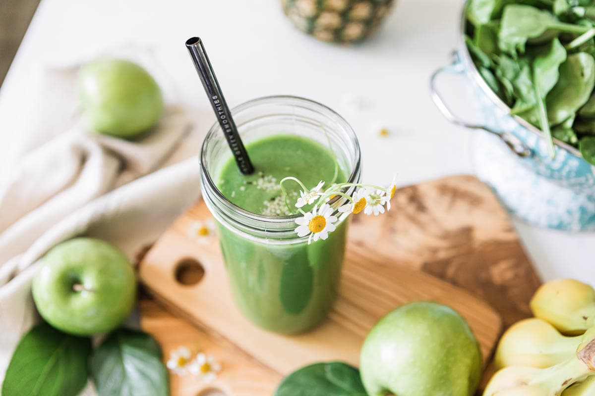 Green apple smoothie with metal straw and floral garnish in a jar, surrounded by fresh green apples, spinach, and bananas.