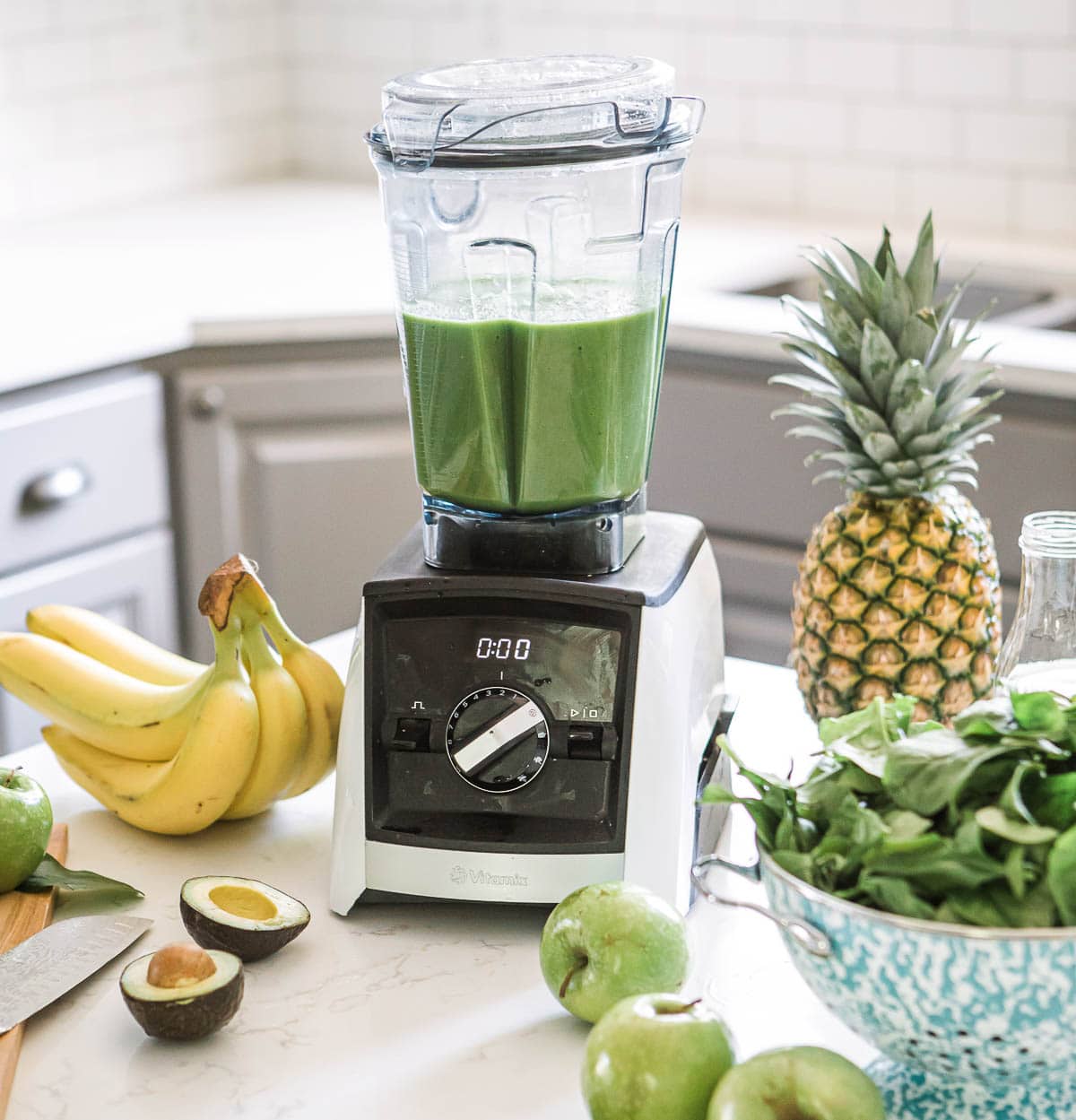 blender full of green smoothie surrounded by fresh produce on a kitchen counter.