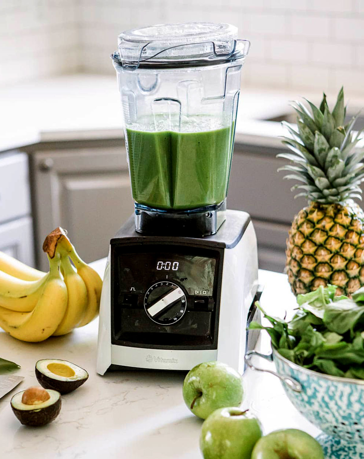 Blender filled with green apple smoothie surrounded by bananas, avocado, pineapple, spinach, and green apples on a kitchen counter.