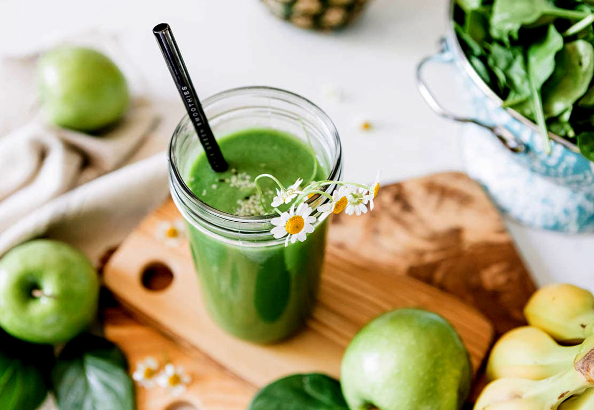 Green apple smoothie with metal straw and floral garnish in a jar, surrounded by fresh green apples, spinach, and bananas.