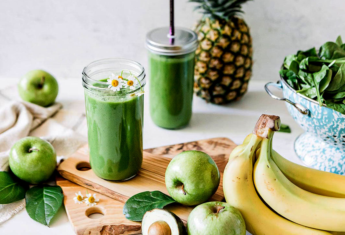 Two jars of green apple smoothie surrounded by fresh spinach, bananas, pineapple, avocado, and green apples and displayed with woodgrain cutting board.