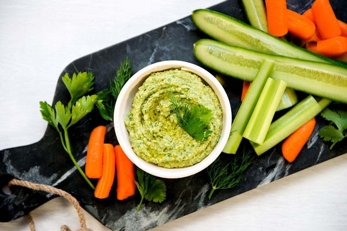 Slate cutting board with bowl of green goddess hummus and fresh cut vegetable sticks.