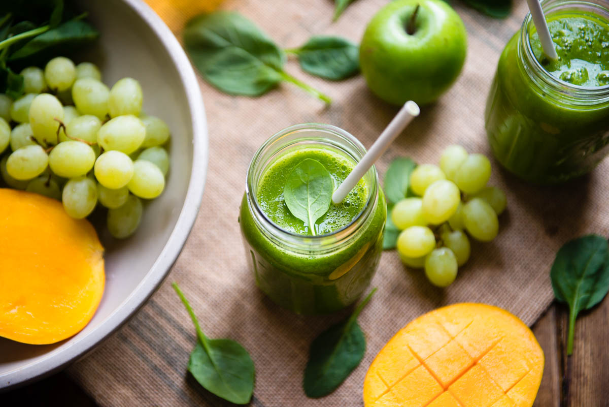 green tea smoothie in a glass mason jar with a paper straw, surrounded by grapes, mango, apple and spinach.