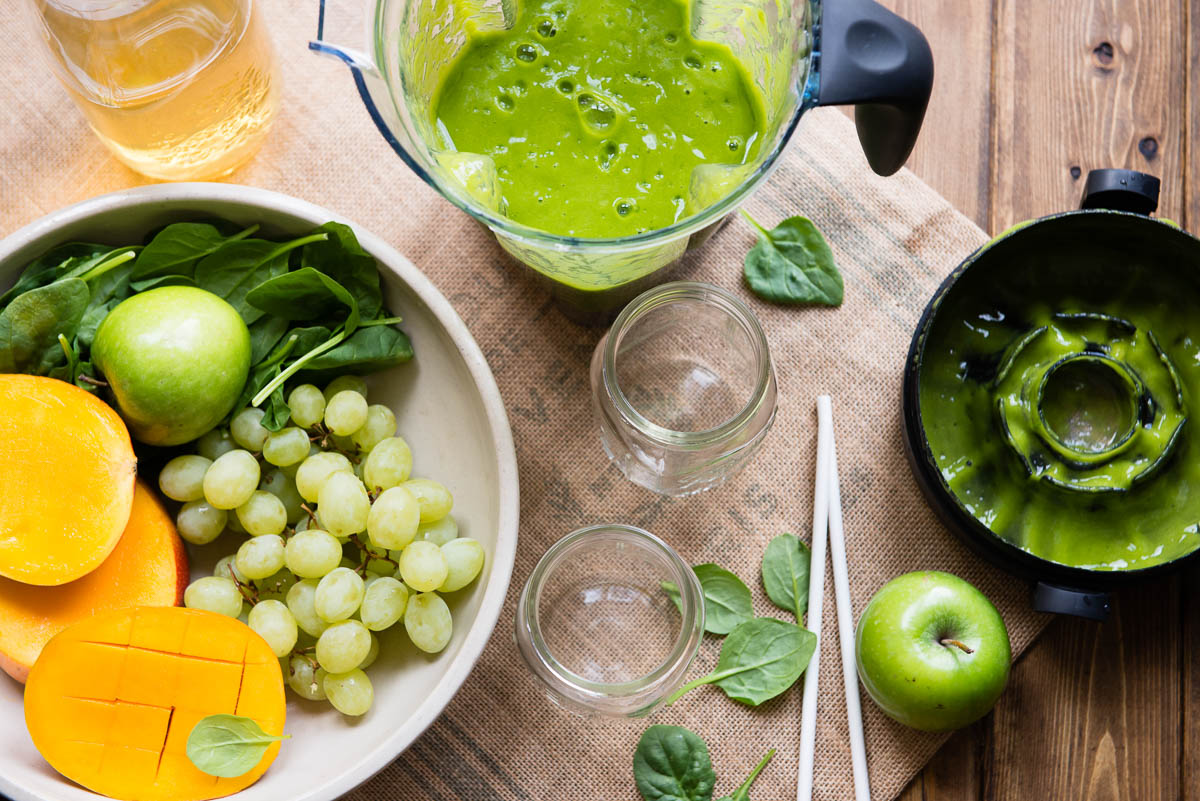 blender container with blended green smoothie next to 2 empty glass jars, 2 white paper straws and a bowl of fresh ingredients.