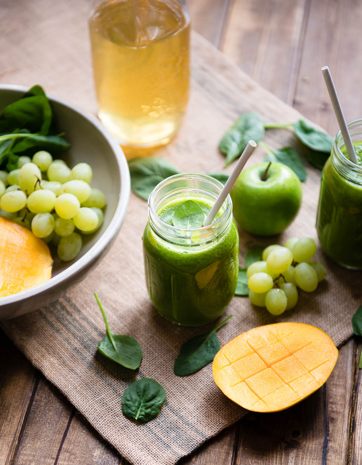 green tea smoothie in a glass mason jar with a paper straw, surrounded by grapes, mango, apple and spinach.