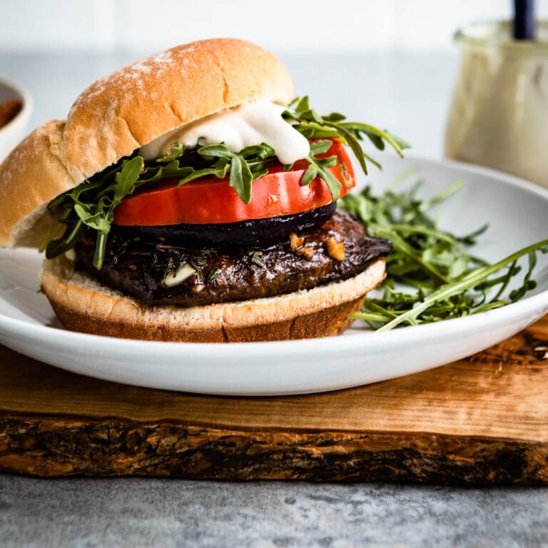 portobello mushroom burger on a white plate topped with arugula, sauce and tomatoes.