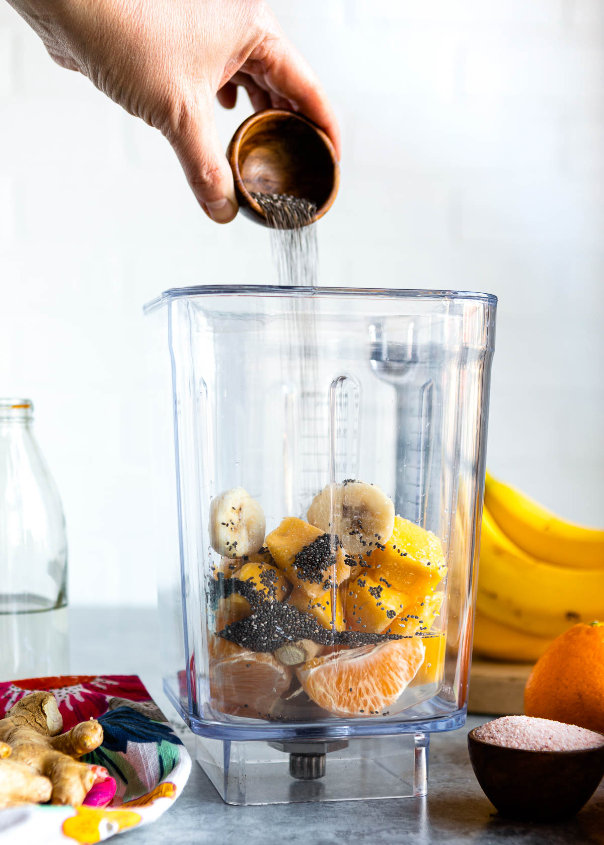 pouring chia seeds into a blender container that containers banana, mango and a tangerine.