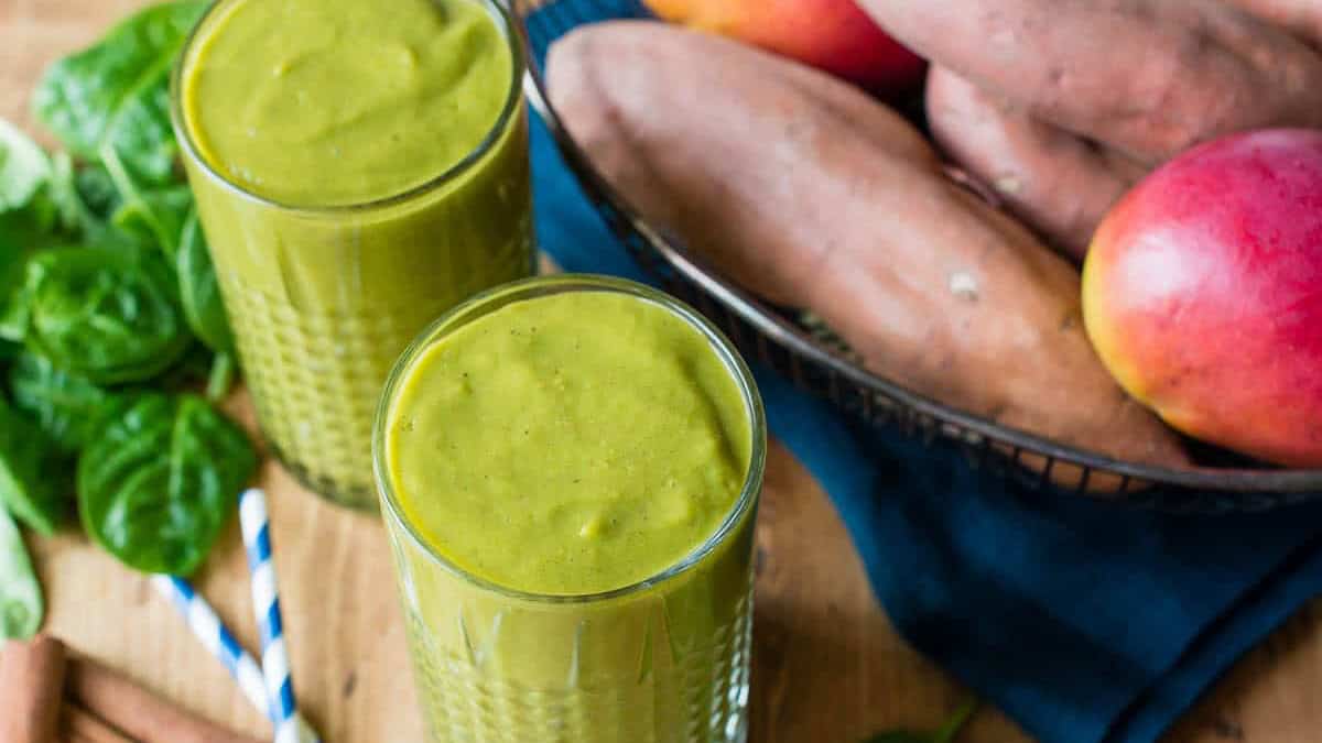 Green smoothie in two glasses with sweet potatoes and apples in background.