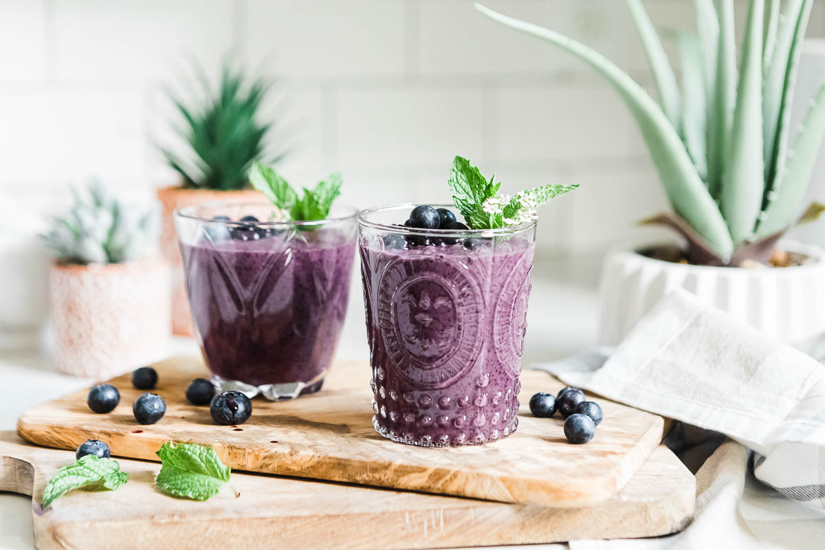 2 glasses full of purple smoothie and topped with fresh fruit and mint sprigs.