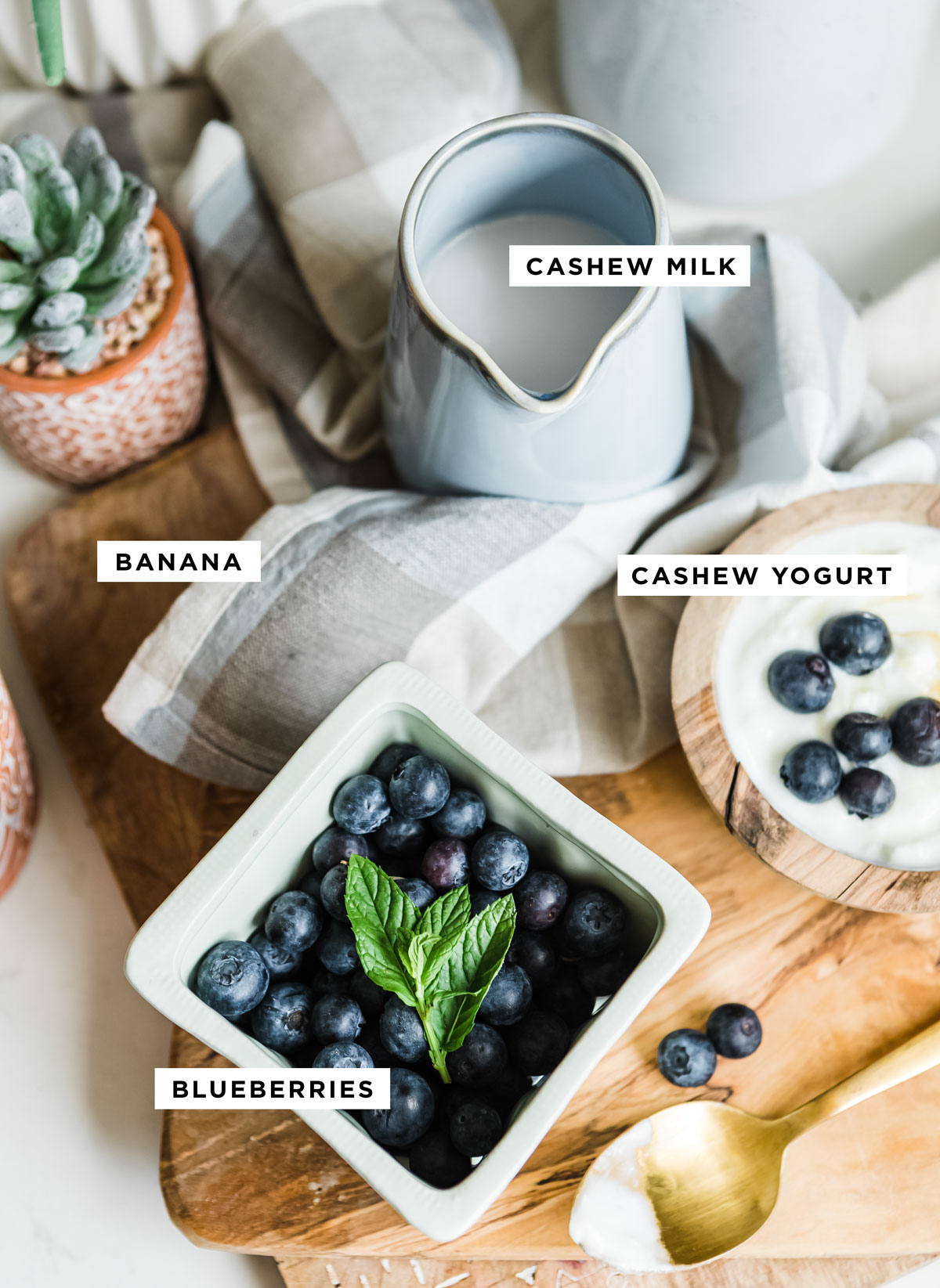 labeled ingredients for healthy smoothie including cashew milk, banana, cashew yogurt and blueberries.