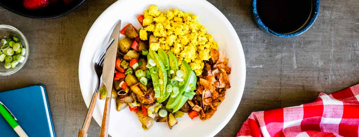 white bowl with cooked breakfast ingredients inside: avocado, potato hash, peppers, eggs