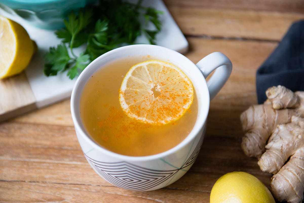 lemon tea with pinch of cayenne pepper in a mug surrounded by cilantro, lemons and ginger.