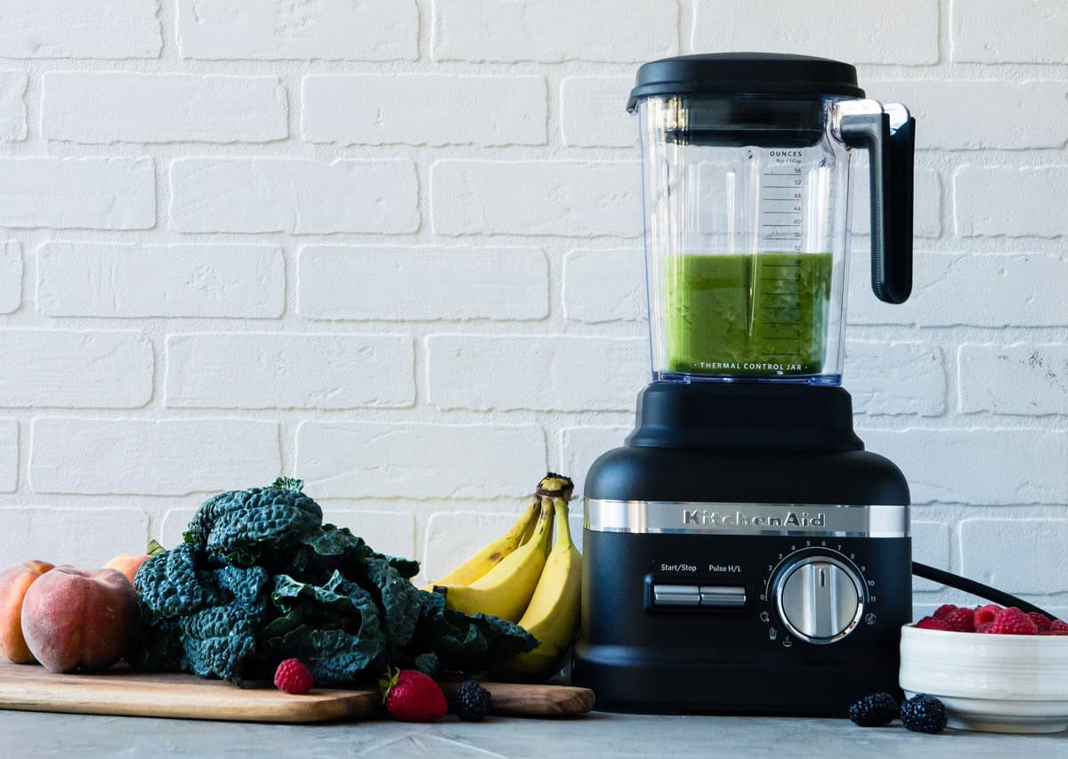 KitchenAid blender with green smoothie and fresh fruits on countertop.