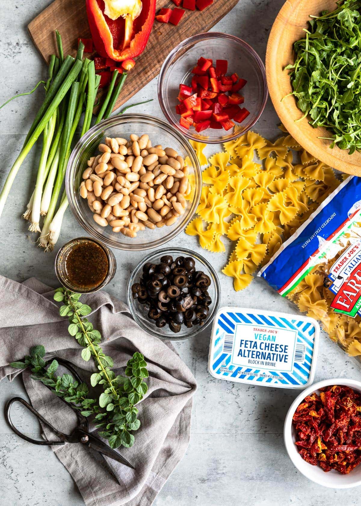 table full of ingredients for healthy lunch including red bell pepper, arugula, white beans, green onions, pasta, black olives, dressing and vegan feta cheese.